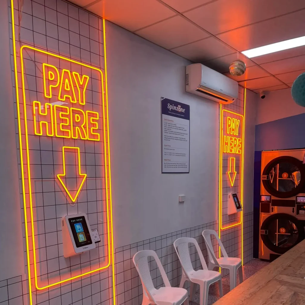 A completed commercial lighting project by Eddy Cullen Electrical at a laundromat in Bowral, featuring a bright orange 'Pay Here' sign. The warm glow improving visibility and clearly guides customers to the payment area.