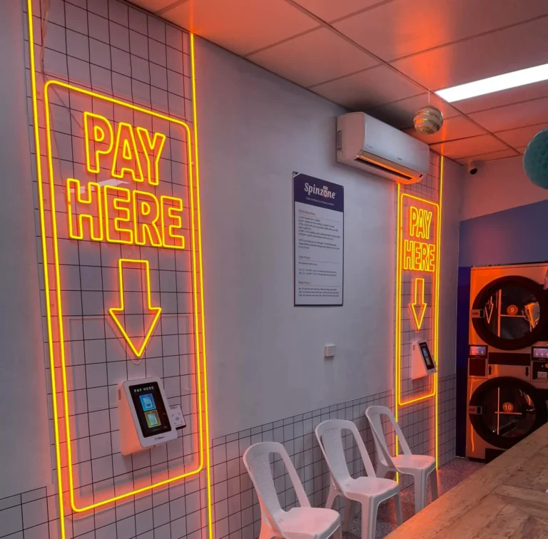 A commercial lighting job completed by Eddy Cullen Electrical in a Tahmoor laundromat, featuring a vibrant orange illuminated ' Pay Here' Sign. The warm orange glow enhances visibility and provides concise directions for all customers.
